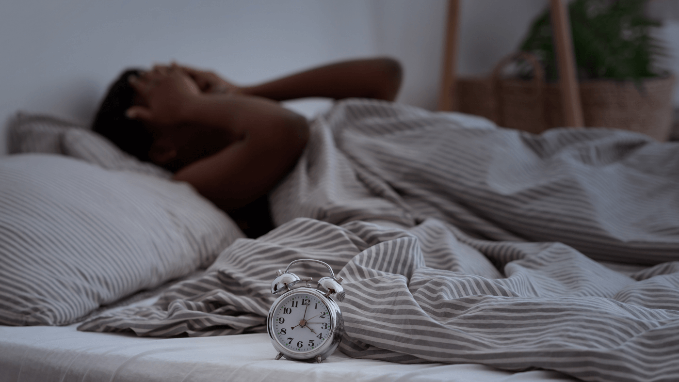 Person lying in bed holding head with alarm clock in foreground